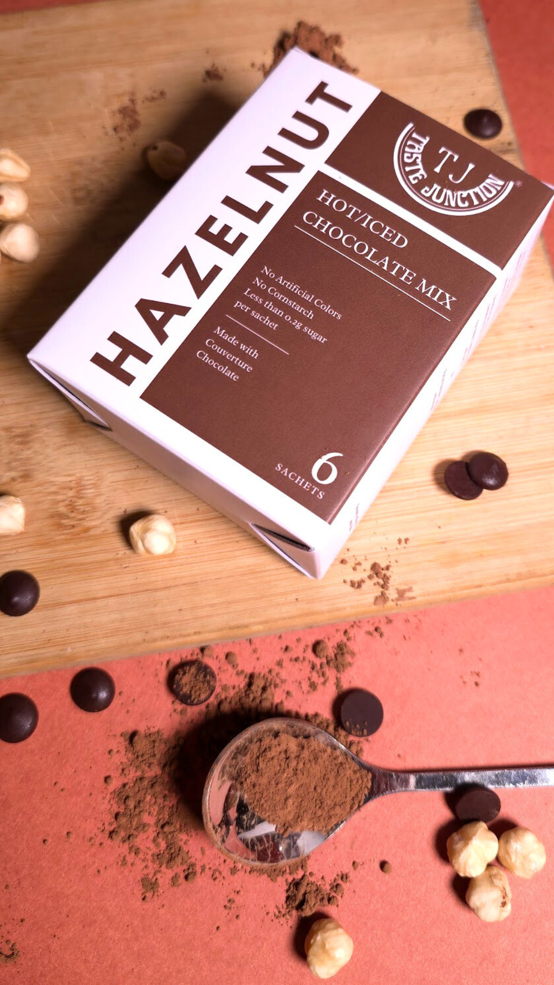 Box of Taste JunctionHazelnut Hot Cocoa Mix on a wooden surface with cocoa powder and hazelnuts.