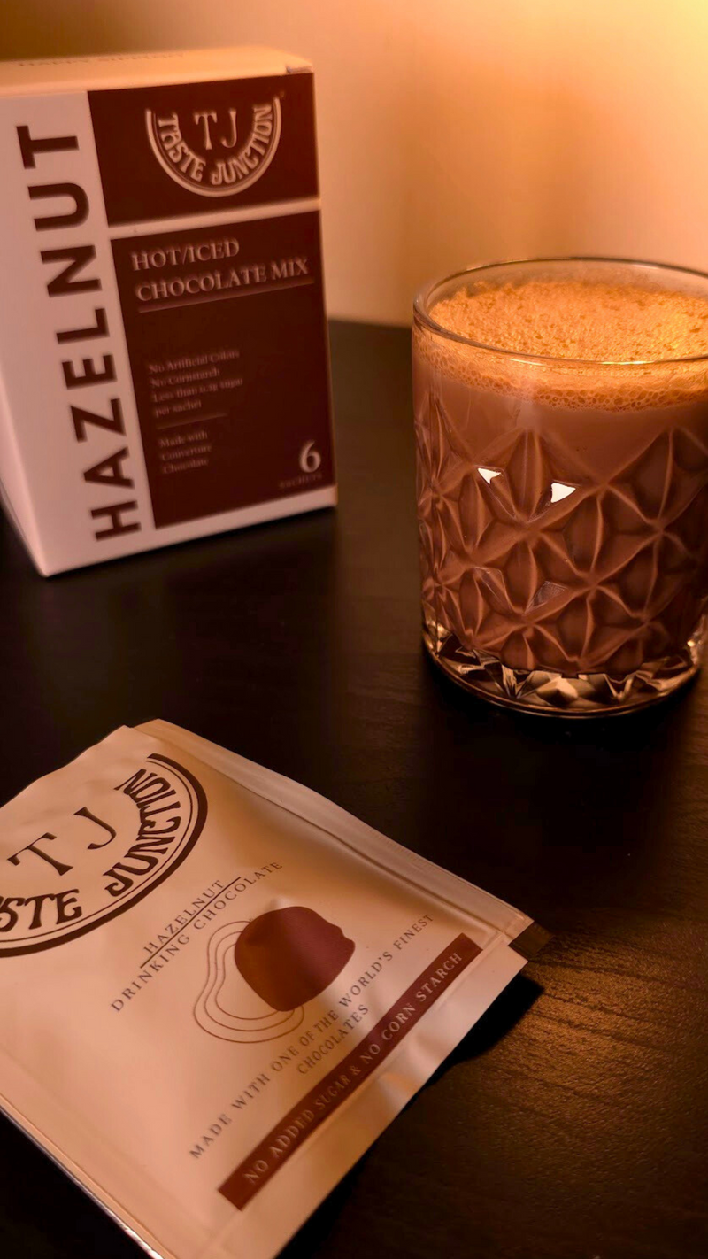 Taste Junction hazelnut hot cocoa mix packaging with a glass of hot cocoa on a wooden surface.