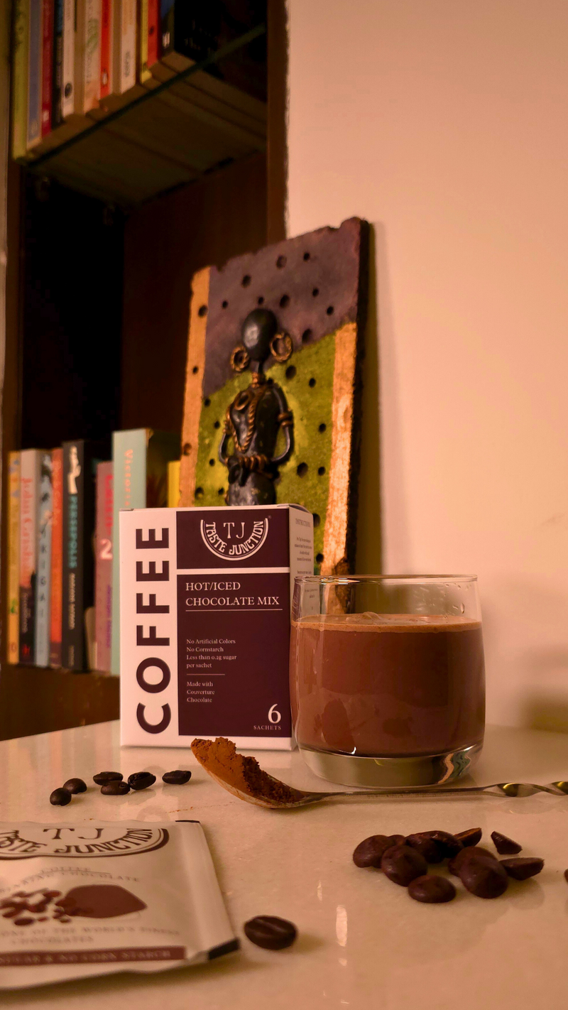Glass of coffee with a box of coffee and coffee beans on a table, with a bookshelf in the background.